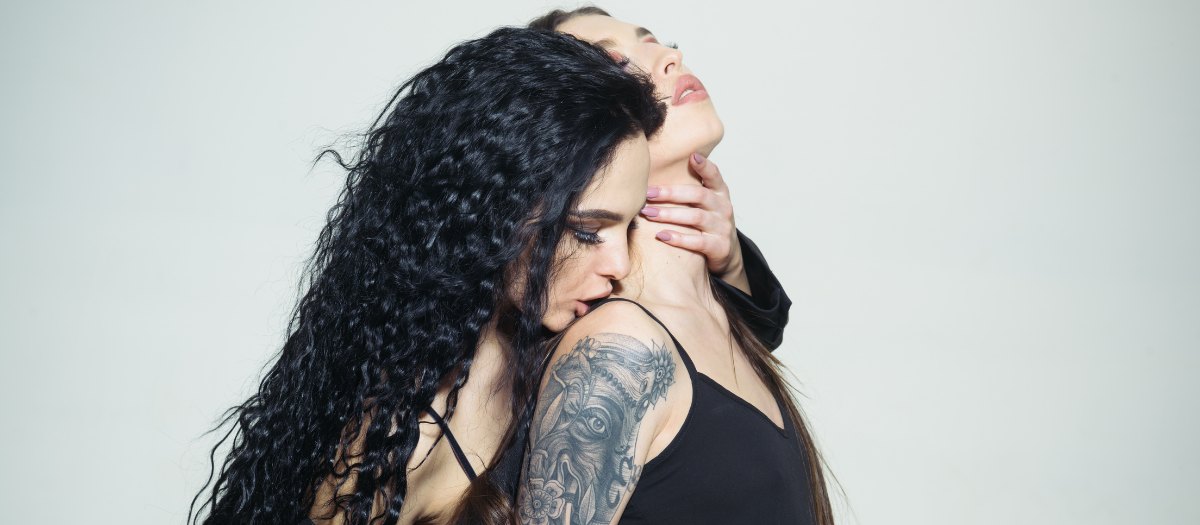 A dark-haired woman with a sleeve tattoo kissing another woman's neck against a plain white wall