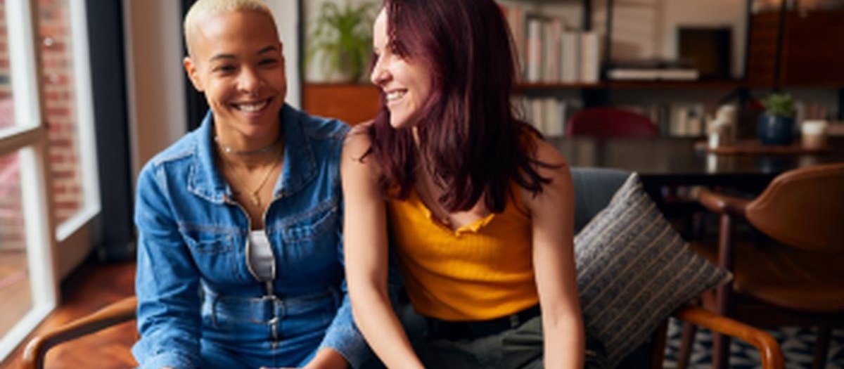 Two women sitting together in a sunlit cafe, mid-conversation and laughing