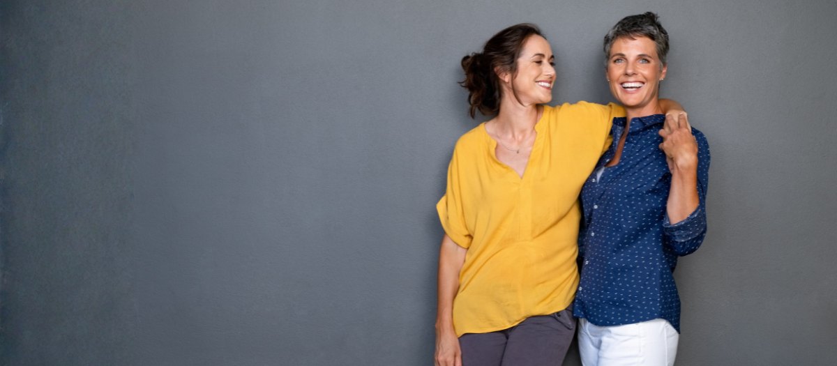Two women standing side by side against a grey wall, one with an arm around the other, both laughing