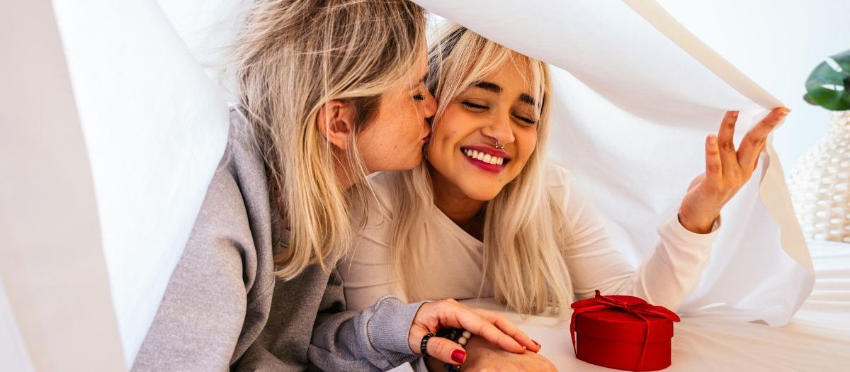 Two women laughing under a white sheet at home, one kissing the other on the cheek
