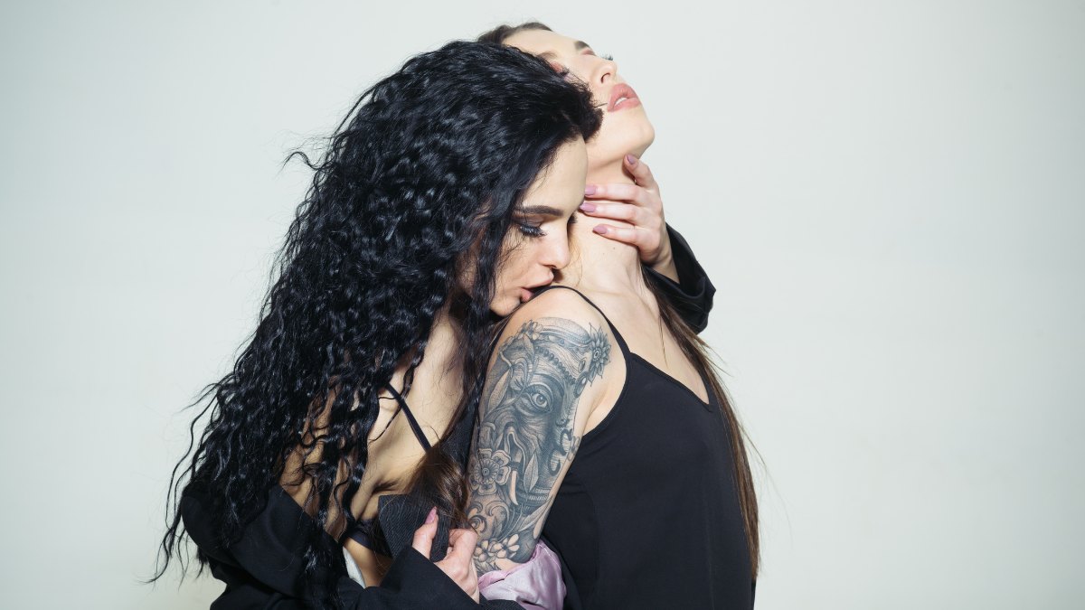 A dark-haired woman with a sleeve tattoo kissing another woman's neck against a plain white wall