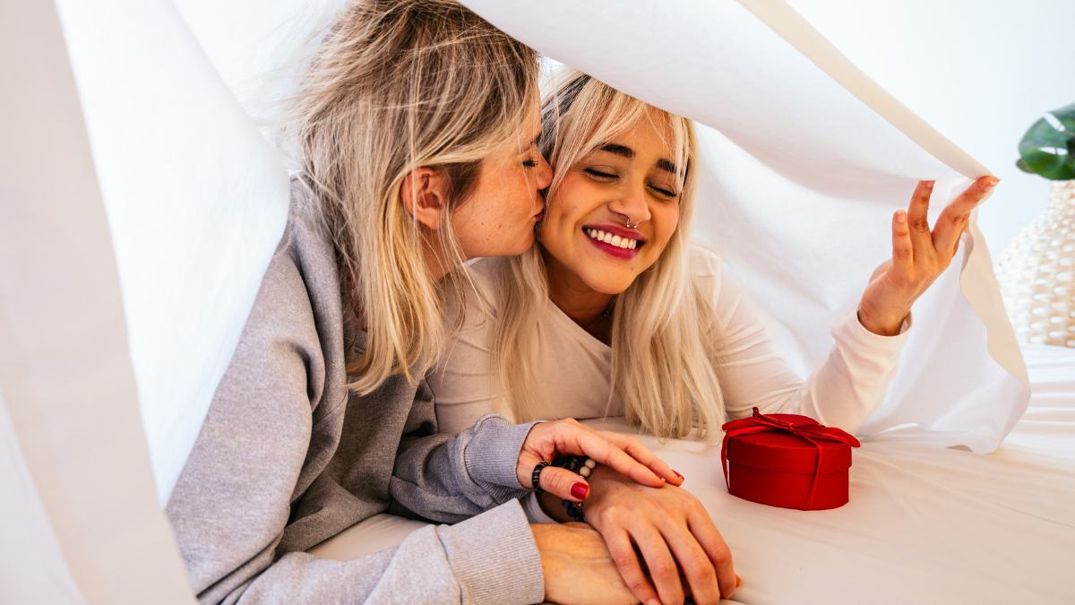 Two women laughing under a white sheet at home, one kissing the other on the cheek