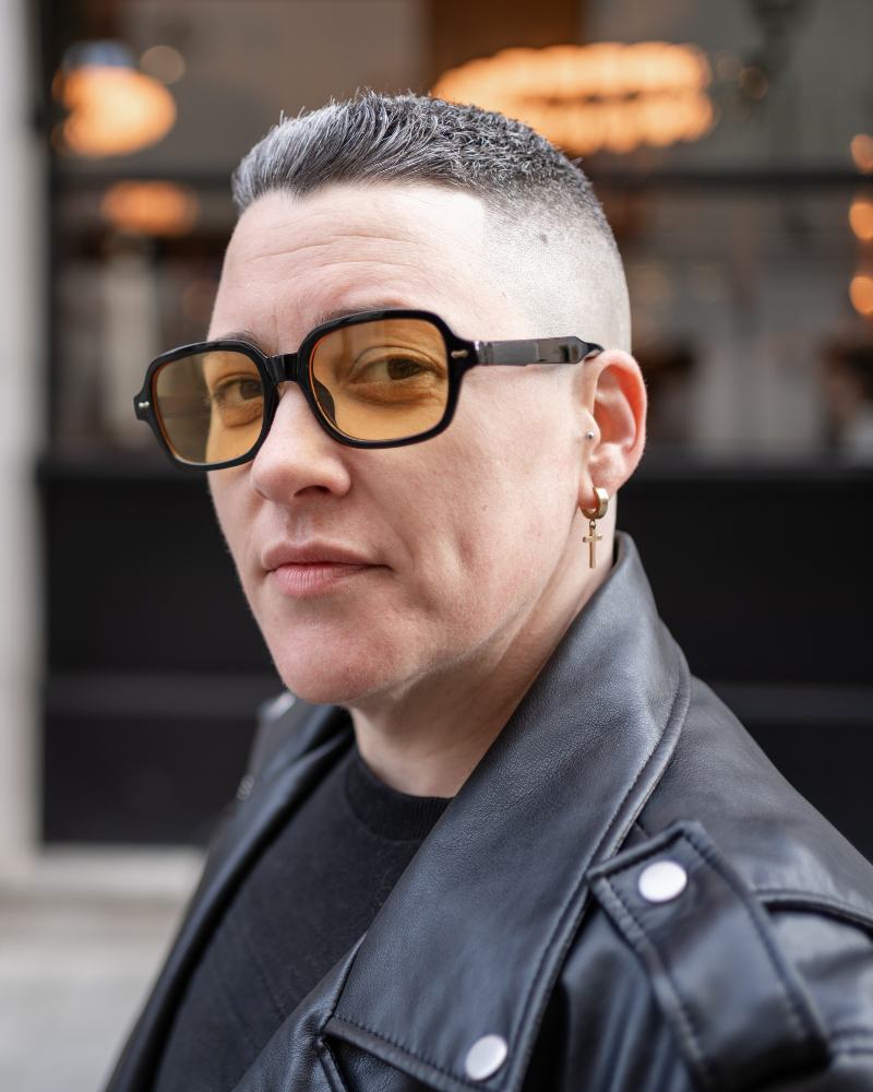 A woman in a leather jacket standing on a city street, looking towards the camera