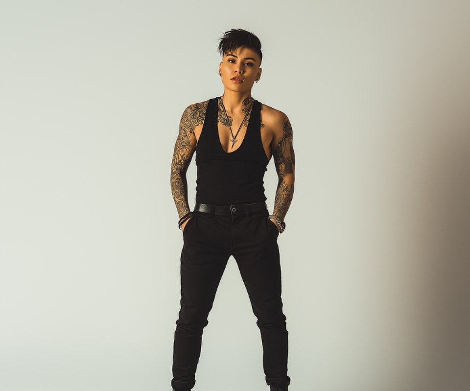 A butch woman standing with hands in pockets, tattooed arms, in a plain studio