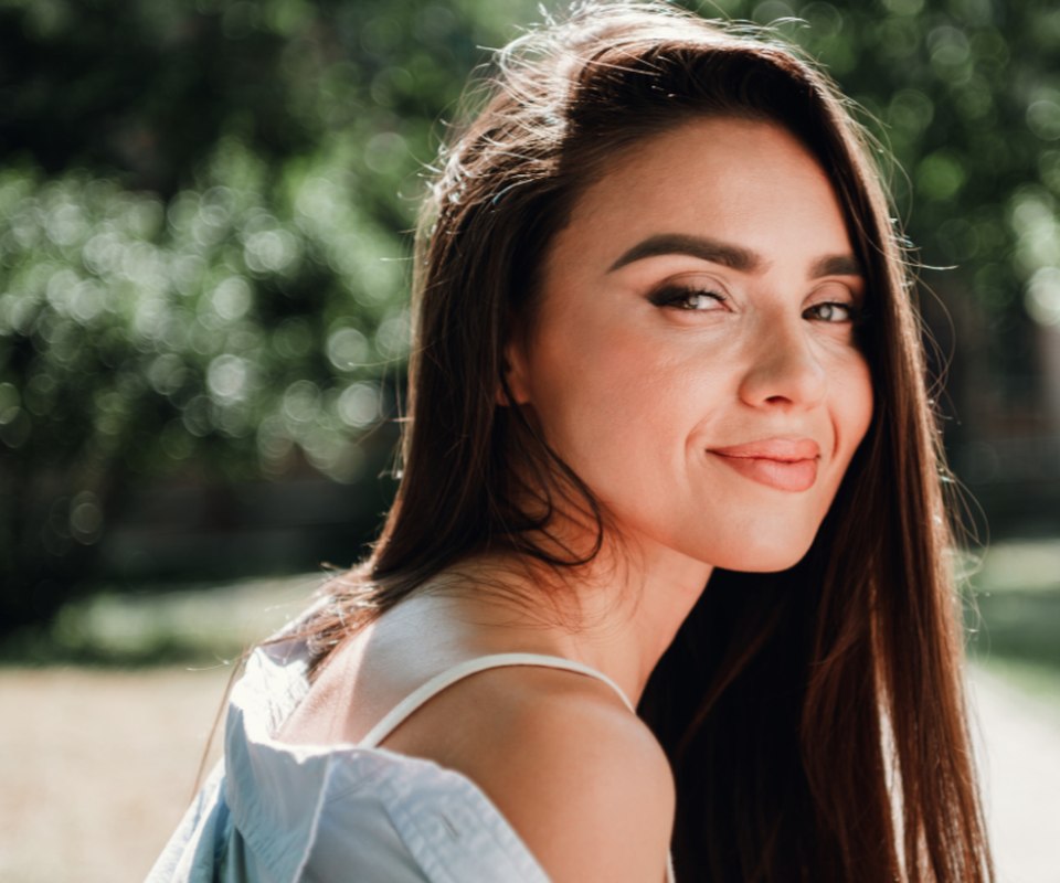 A woman with long dark hair smiling over her shoulder outdoors in daylight