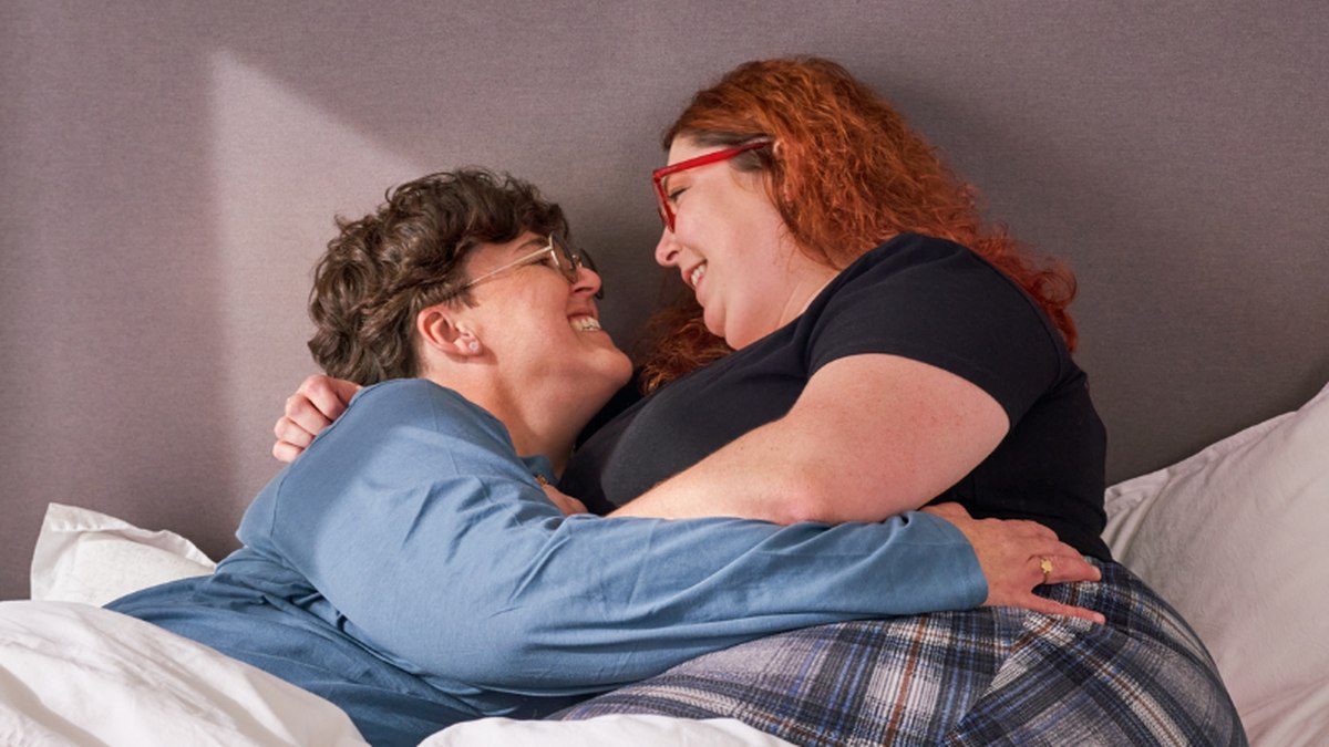 Two women laughing together in bed, one resting an arm around the other